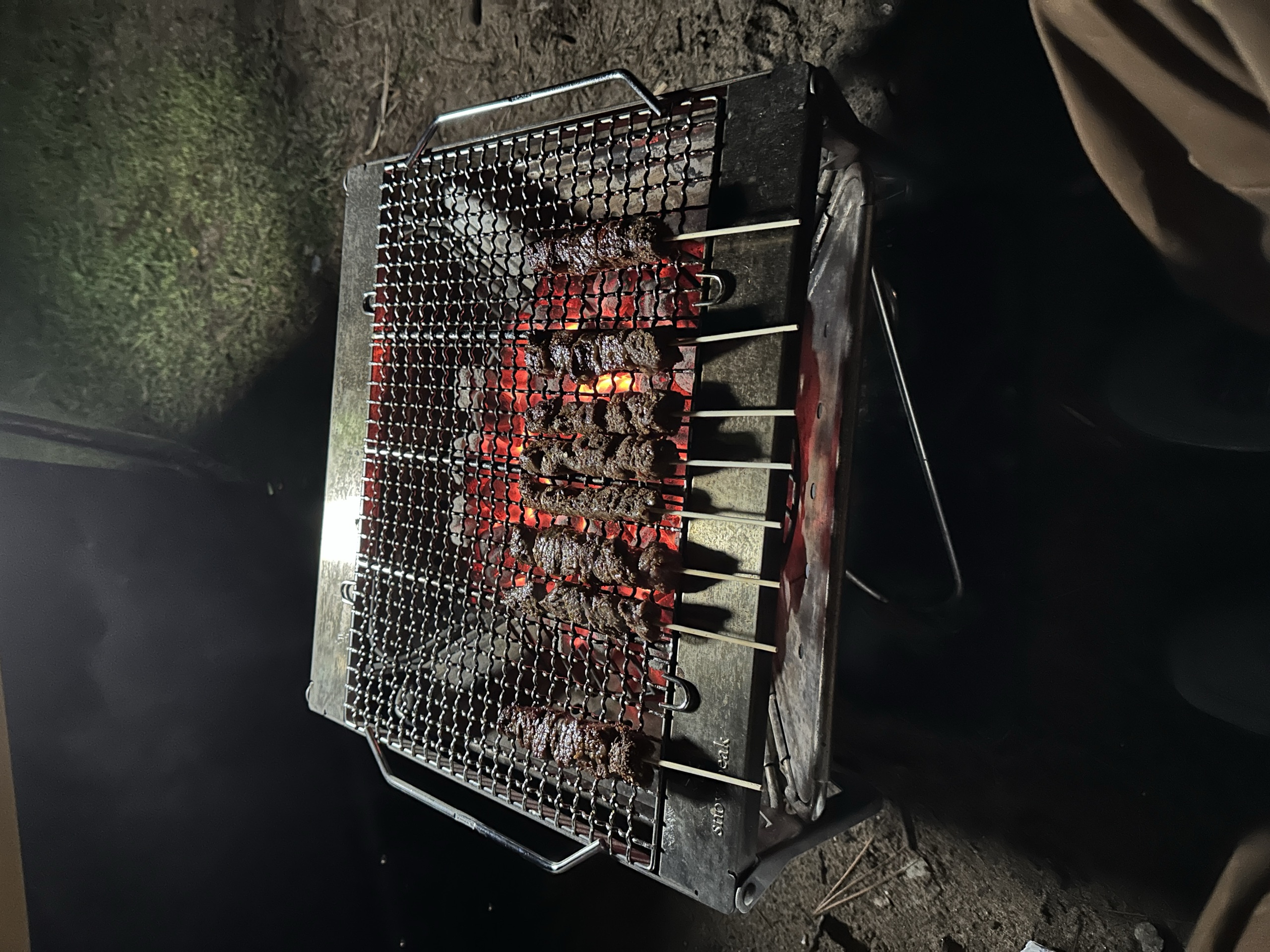 Yakitori skewers cooking on a small charcoal grill at night, the meat browning over the coals