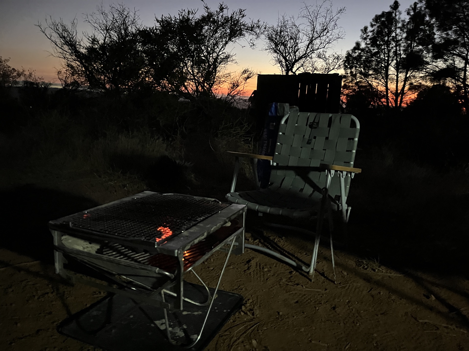 A portable BBQ grill glowing red with coals at dusk, a folding chair beside it on a mountain clearing
