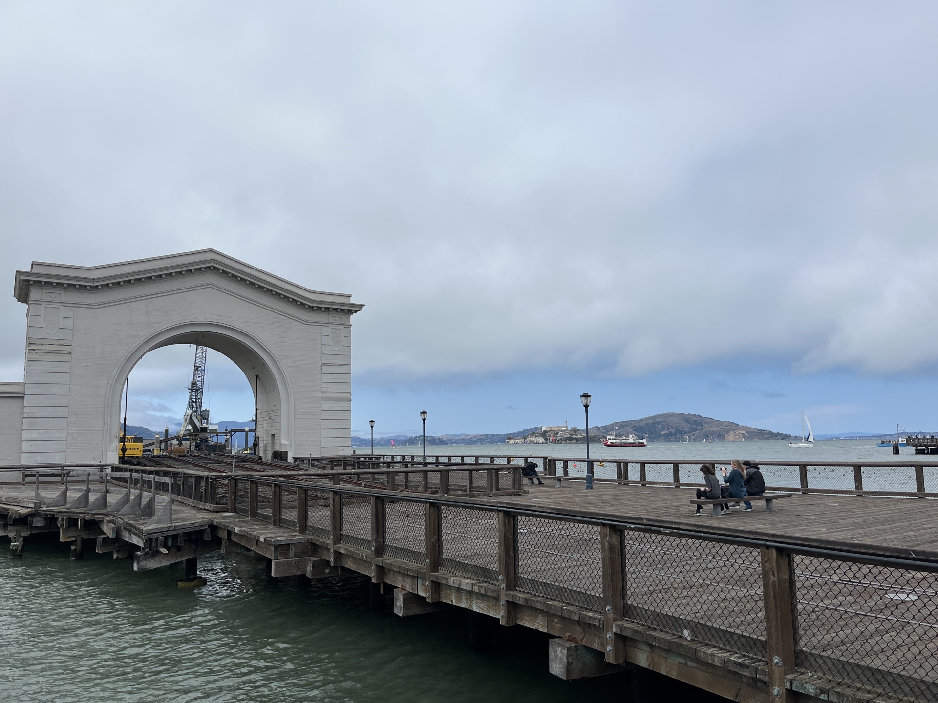 A white stone arch on a wooden pier, Alcatraz Island visible across the water in the background